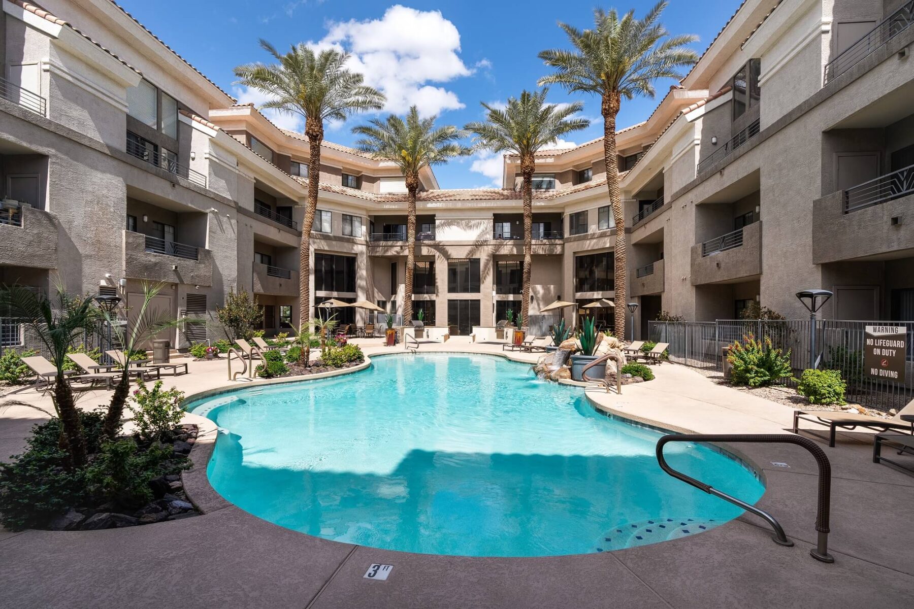 A courtyard swimming pool surrounded by lounge chairs, palm trees, and three-story beige apartment buildings under a blue sky with scattered clouds.