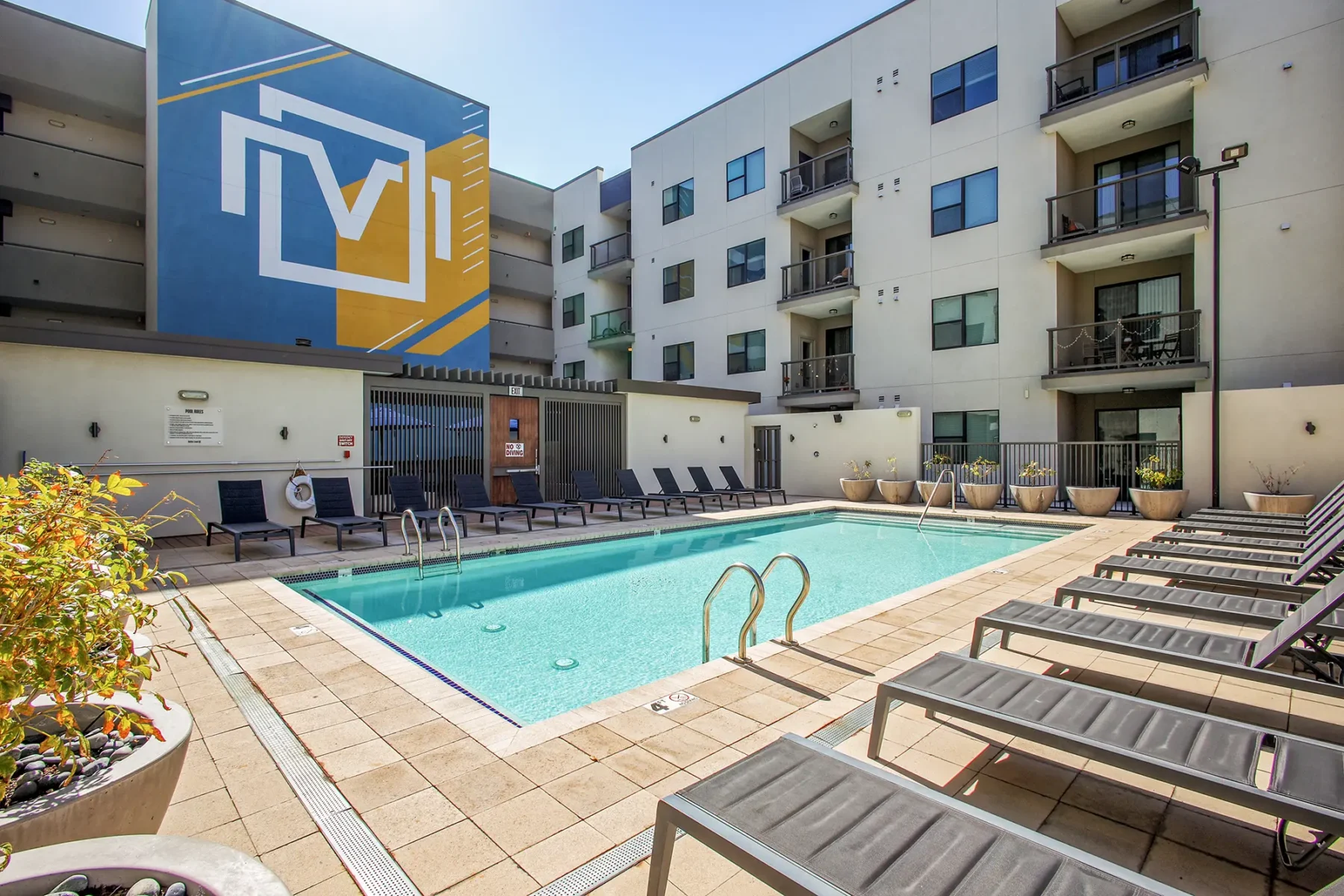 Modern apartment complex courtyard with a rectangular swimming pool, lounge chairs, potted plants, and balconies. A large colorful mural decorates one building wall on a sunny day.