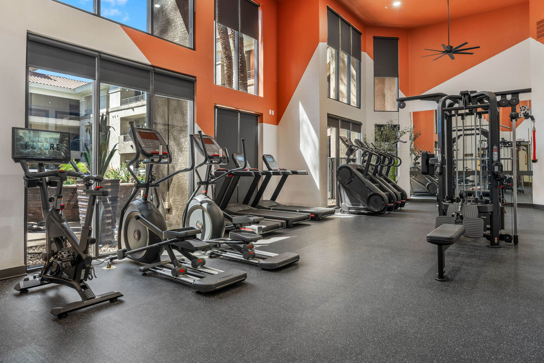Modern gym with cardio machines, treadmills, stationary bikes, and a weight machine. Large windows let in natural light; orange accent walls and high ceilings create an open, airy atmosphere.