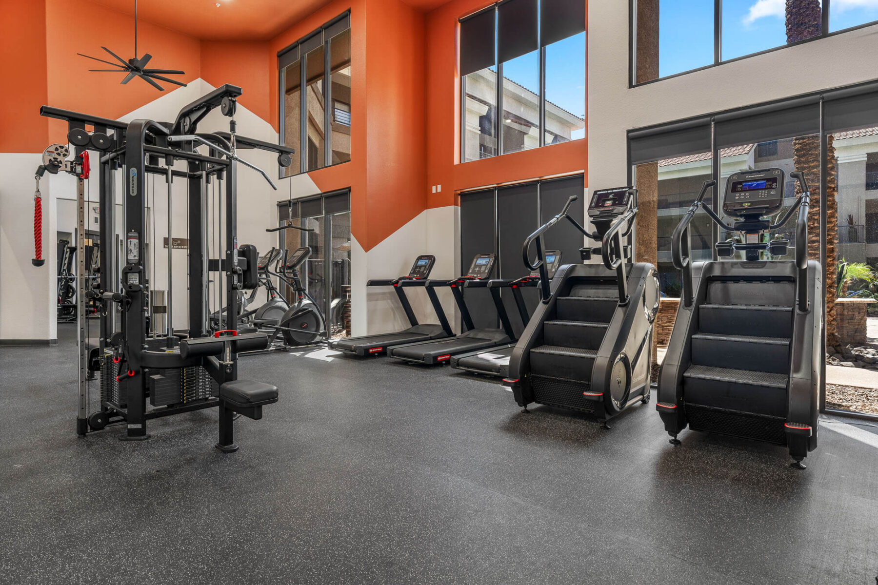 Modern gym with weight machines, treadmills, stair climbers, and large windows letting in natural light. Orange and white walls, rubber flooring, and views of an outdoor area are visible.