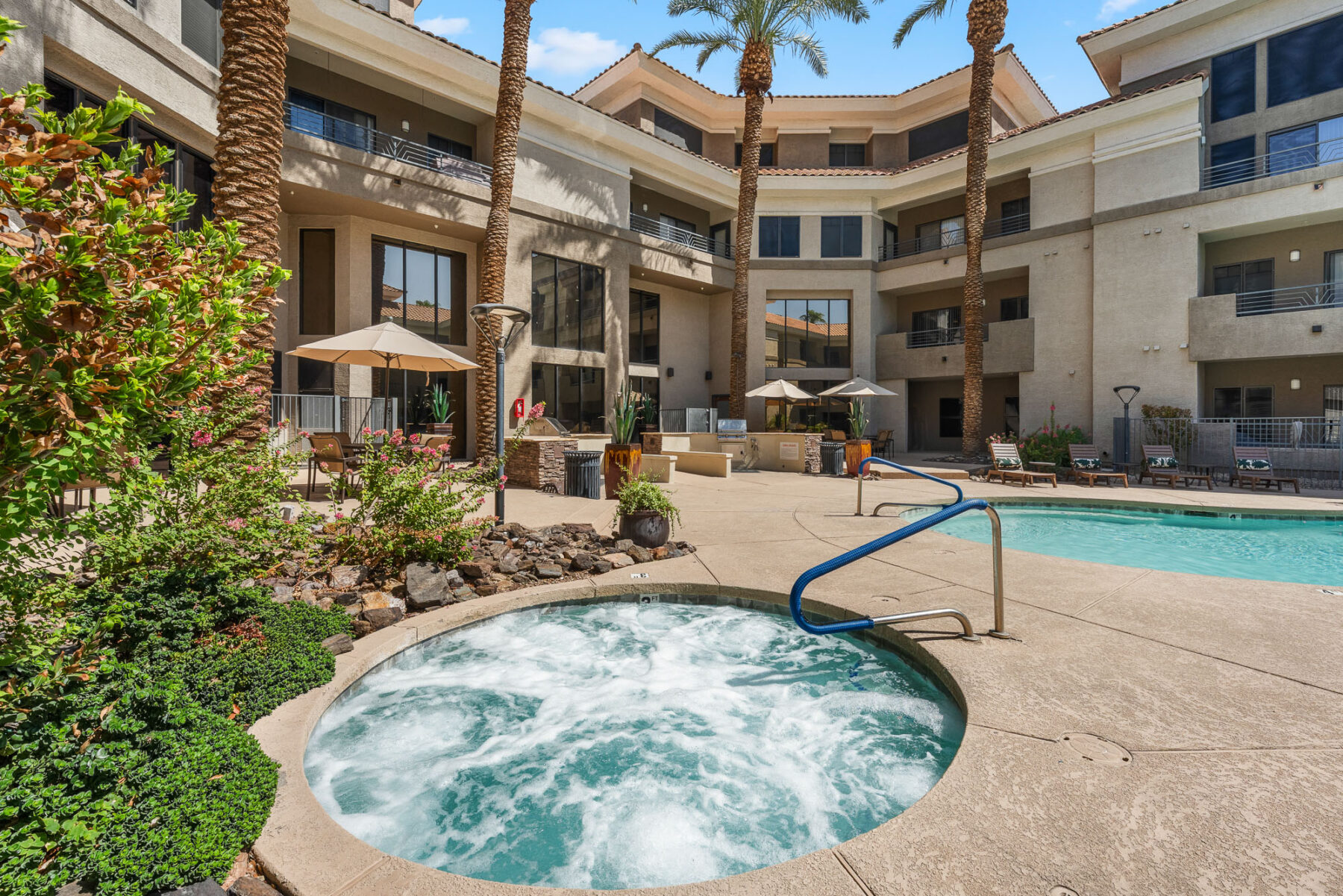 Experience ultimate relaxation at these apartments in Salem, OR, featuring an outdoor area with a bubbling hot tub in the foreground, a sparkling swimming pool, lounge chairs, umbrellas, and swaying palm trees—all set against the backdrop of a three-story beige apartment building under a clear blue sky.