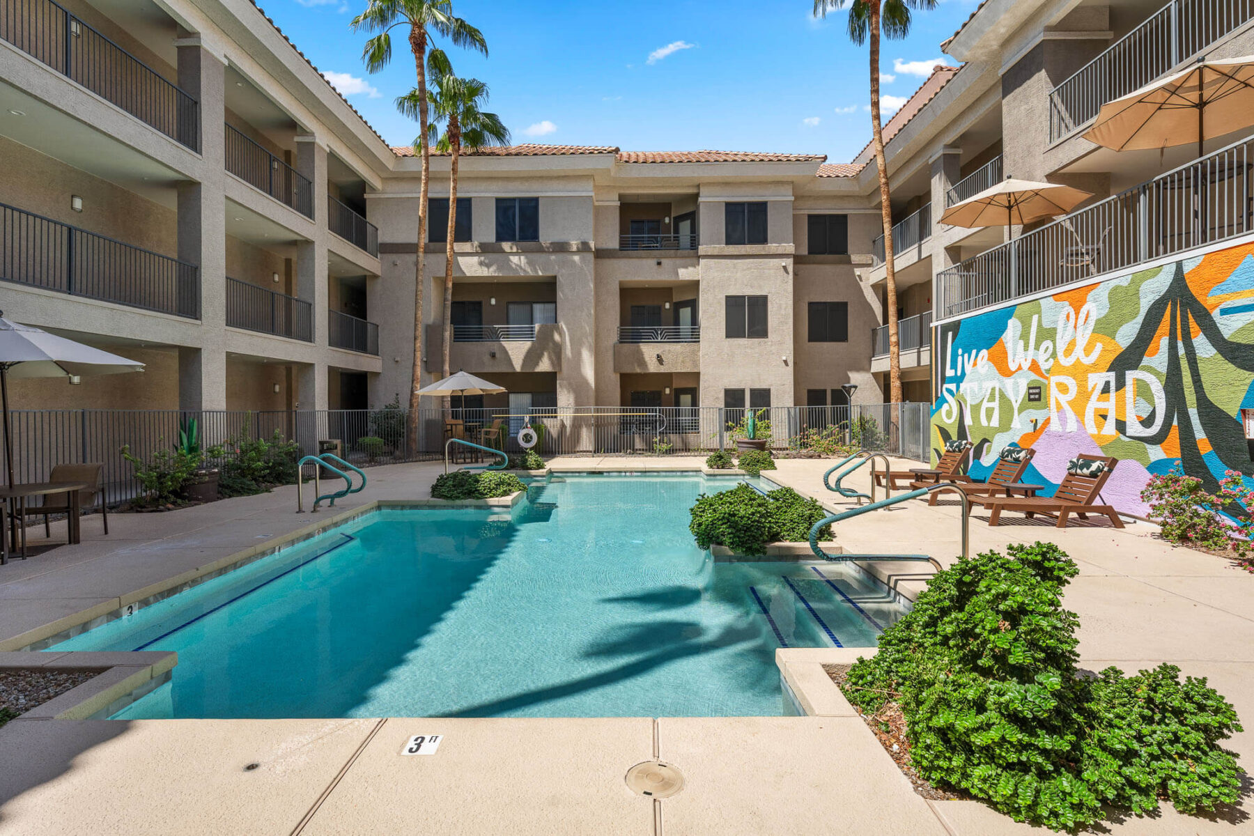 Apartment courtyard with a rectangular pool, lounge chairs, umbrellas, and palm trees. A colorful mural on one wall reads Live Well Stay Rad. Three-story building surrounds the pool area under a clear blue sky.